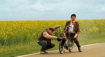 Movie still from “Mr. Bean's Holiday” (2007), directed by Steve Bendelack – A man kneeling down next to a man riding a bike; Wide shot, Low angle