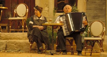 Movie still from “Mr. Bean's Holiday” (2007), directed by Steve Bendelack – An older man and woman sitting at an outdoor table with an accordion; Wide shot, High angle