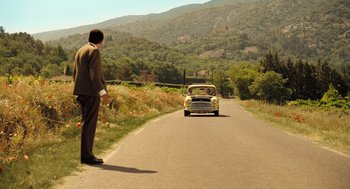 Movie still from “Mr. Bean's Holiday” (2007), directed by Steve Bendelack – A man standing on the side of a road next to a car; Extreme Wide shot, High angle