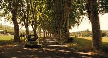 An old yellow car driving down a tree lined road