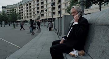 Movie still from “Mr. Bean's Holiday” (2007), directed by Steve Bendelack – An older man sitting on a bench talking on a cell phone; Medium shot, Low angle