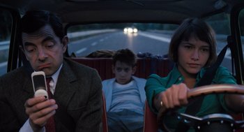 Movie still from “Mr. Bean's Holiday” (2007), directed by Steve Bendelack – Two people and a child in the back seat of a car; Medium shot, Over the shoulder angle