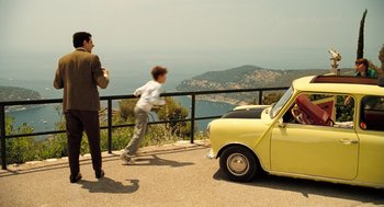 Movie still from “Mr. Bean's Holiday” (2007), directed by Steve Bendelack – A man standing next to a yellow car on the side of a road; Wide shot, High angle