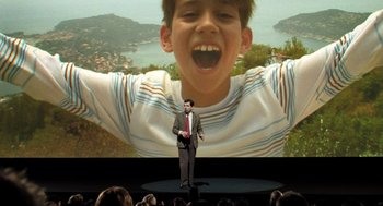 Movie still from “Mr. Bean's Holiday” (2007), directed by Steve Bendelack – A man standing in front of a large picture of a boy; Medium shot, High angle