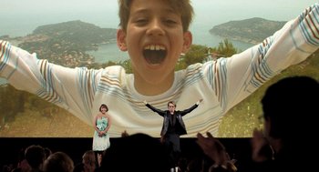 Movie still from “Mr. Bean's Holiday” (2007), directed by Steve Bendelack – A man and a woman standing in front of a large screen; Wide shot, Low angle