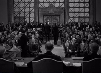 Movie still from “Mr. Deeds Goes to Town” (1936), directed by Frank Capra – An audience is sitting in front of a man in a black suit; Wide shot, High angle