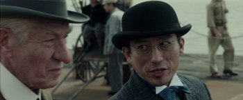 Movie still from “Mr. Holmes” (2015), directed by Bill Condon – A man wearing a hat and glasses with other people in the background; Close Up shot, Over the shoulder angle