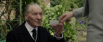 Movie still from “Mr. Holmes” (2015), directed by Bill Condon – An older man in a suit holding a business card; Close Up shot, Over the shoulder angle