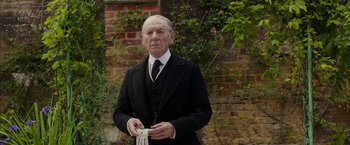 Movie still from “Mr. Holmes” (2015), directed by Bill Condon – An older man in a suit and tie standing in front of a brick wall; Medium shot, Low angle