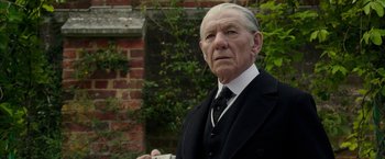 Movie still from “Mr. Holmes” (2015), directed by Bill Condon – An older man in a suit and tie standing in front of a brick wall; Close Up shot, Low angle
