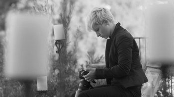 Movie still from “Much Ado About Nothing” (2012), directed by Joss Whedon – A person sitting down holding a camera and looking at the camera; Medium shot, High angle