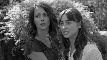 Movie still from “Much Ado About Nothing” (2012), directed by Joss Whedon – Two young women standing next to each other in front of bushes; Close Up shot, Over the shoulder angle