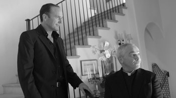 Movie still from “Much Ado About Nothing” (2012), directed by Joss Whedon – A priest and a man standing next to each other in front of a staircase; Medium shot, Low angle
