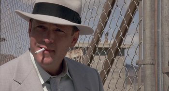 Movie still from “Mulholland Falls” (1996), directed by Lee Tamahori – A man wearing a fedora smoking a cigarette in front of a chain link fence; Close Up shot, Low angle