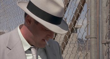 Movie still from “Mulholland Falls” (1996), directed by Lee Tamahori – A man wearing a hat and a suit smoking a cigarette in front of a chain link fence; Close Up shot, Low angle