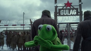 Movie still from “Muppets Most Wanted” (2014), directed by James Bobin – A green stuffed animal sitting in front of a fence; Medium shot, Over the shoulder angle
