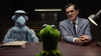 Movie still from “Muppets Most Wanted” (2014), directed by James Bobin – A man sitting at a table next to a stuffed animal; Medium shot, Over the shoulder angle
