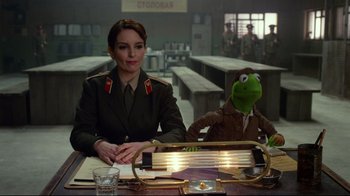 Movie still from “Muppets Most Wanted” (2014), directed by James Bobin – A woman in a uniform sitting at a table next to a muppet; Medium shot, Over the shoulder angle
