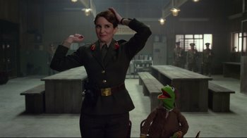 Movie still from “Muppets Most Wanted” (2014), directed by James Bobin – A woman in a military uniform standing next to a man in a kermit costume; Medium shot, Over the shoulder angle