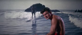Movie still from “Muscle Beach Party” (1964), directed by William Asher – A man standing in the water with a surfboard in front of him; Medium shot, Low angle