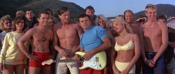 Movie still from “Muscle Beach Party” (1964), directed by William Asher – A man holding a guitar in front of a group of people; Medium shot, High angle