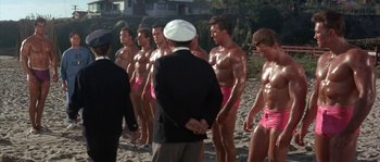 Movie still from “Muscle Beach Party” (1964), directed by William Asher – A group of shirtless men standing next to each other on a beach; Wide shot, High angle