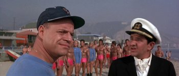 Movie still from “Muscle Beach Party” (1964), directed by William Asher – A group of men in swim trunks standing next to each other on a beach; Medium shot, Over the shoulder angle