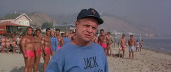 Movie still from “Muscle Beach Party” (1964), directed by William Asher – A man standing on a beach with a bunch of men; Medium shot, Over the shoulder angle