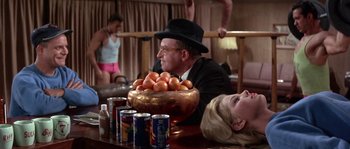Movie still from “Muscle Beach Party” (1964), directed by William Asher – A man in a suit and a hat is sitting at a table with a bowl of oranges; Medium shot, Low angle