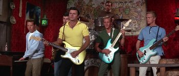 Movie still from “Muscle Beach Party” (1964), directed by William Asher – A group of men are playing guitars in front of a band; Medium shot, Low angle