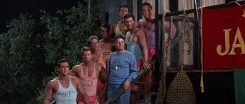 Movie still from “Muscle Beach Party” (1964), directed by William Asher – A group of shirtless men standing next to each other; Wide shot, High angle