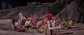 Movie still from “Muscle Beach Party” (1964), directed by William Asher – A group of people sitting on the beach watching a man stand up; Wide shot, High angle