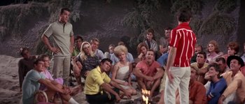Movie still from “Muscle Beach Party” (1964), directed by William Asher – A group of people sitting on the ground near a fire pit; Wide shot, High angle