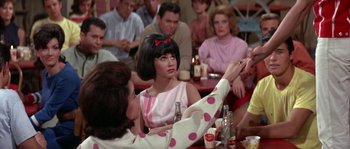 Movie still from “Muscle Beach Party” (1964), directed by William Asher – A woman sitting in front of a group of people at a table; Medium shot, Over the shoulder angle