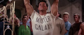Movie still from “Muscle Beach Party” (1964), directed by William Asher – A man in a t - shirt that says " mister galaxy "; Medium shot, Low angle