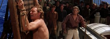 Movie still from “Mutiny on the Bounty” (1962), directed by George Seaton – A group of people standing around a man with a naked torso; Medium shot, Over the shoulder angle