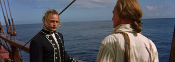 Movie still from “Mutiny on the Bounty” (1962), directed by George Seaton – Two men on a boat in front of a body of water; Medium shot, Over the shoulder angle