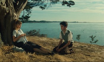 Movie still from “My American Uncle” (1980), directed by Alain Resnais – Two people sitting on the ground near a body of water; Wide shot, Low angle