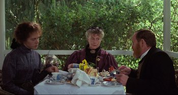 Movie still from “My Brilliant Career” (1979), directed by Gillian Armstrong – An older woman sitting at a table with a plate of hot dogs; Medium shot, Over the shoulder angle