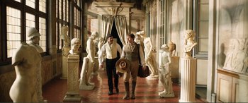 Movie still from “My Cousin Rachel” (2017), directed by Roger Michell – A couple of men walking down a hall way with statues; Wide shot, Over the shoulder angle
