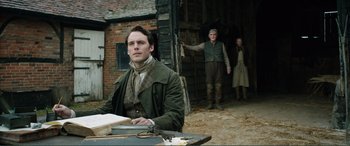 Movie still from “My Cousin Rachel” (2017), directed by Roger Michell – A man sitting at a table in front of a barn; Medium shot, Over the shoulder angle