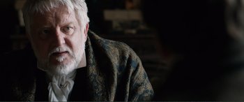 Movie still from “My Cousin Rachel” (2017), directed by Roger Michell – An older man with a white beard wearing a black and gold jacket; Close Up shot, Over the shoulder angle