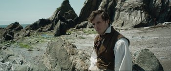 Movie still from “My Cousin Rachel” (2017), directed by Roger Michell – A man in a vest and tie standing next to rocks; Medium shot, Over the shoulder angle