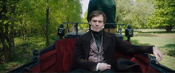 Movie still from “My Cousin Rachel” (2017), directed by Roger Michell – A man sitting on a red couch in a park; Medium shot, Over the shoulder angle