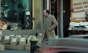 Movie still from “My Dinner with Andre” (1981), directed by Louis Malle – A man walking down the street in a trench coat; Wide shot, High angle