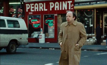 Movie still from “My Dinner with Andre” (1981), directed by Louis Malle – A man is walking down the street wearing a brown coat; Wide shot, Over the shoulder angle