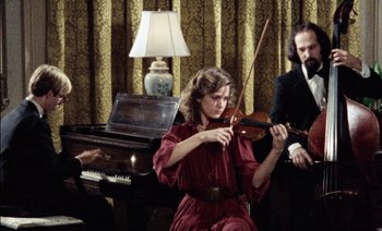 Movie still from “My Dinner with Andre” (1981), directed by Louis Malle – A woman in a red dress playing a violin in front of two other people; Medium shot, High angle