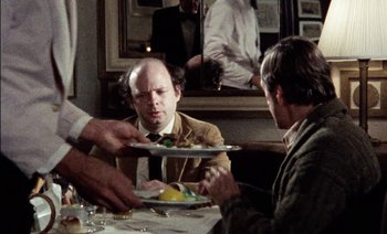 Movie still from “My Dinner with Andre” (1981), directed by Louis Malle – A man sitting at a table with a plate of food on it; Medium shot, Over the shoulder angle