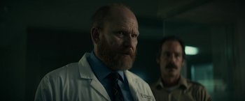 Movie still from “Fractured” (2019), directed by Brad Anderson – A man in a lab coat standing next to another man in a dark room; Close Up shot, Low angle