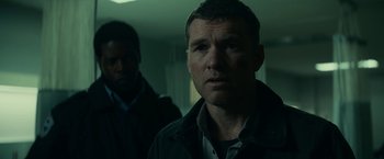 Movie still from “Fractured” (2019), directed by Brad Anderson – A man standing next to another man in a dark room; Close Up shot, Over the shoulder angle